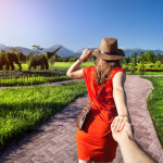 Woman in orange dress and hat holding man by hand and going to Topiary Garden with elephants